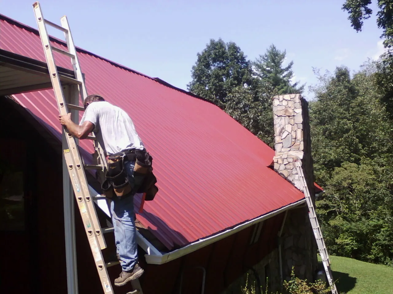 Roofer on a ladder beside a red metal roof and stone chimney — Soffit & Fascia Repair by Roofing Brothers in Veteran